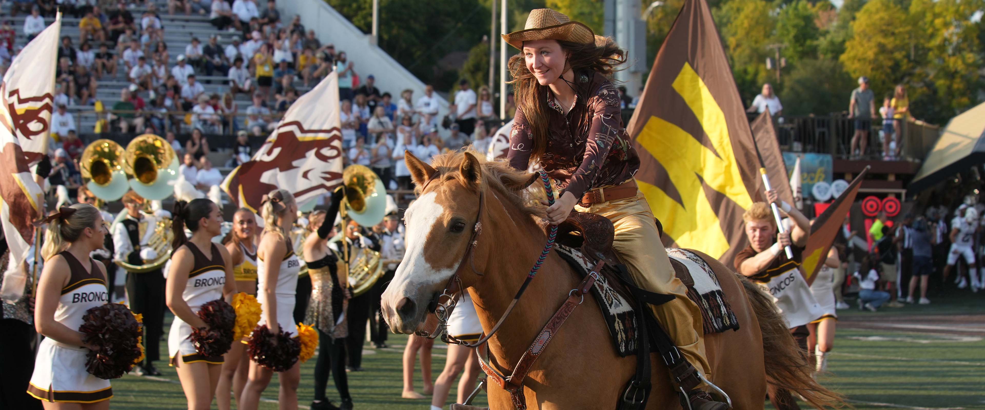 A student riding a Bronco into Waldo Stadium as the band performs on the field.