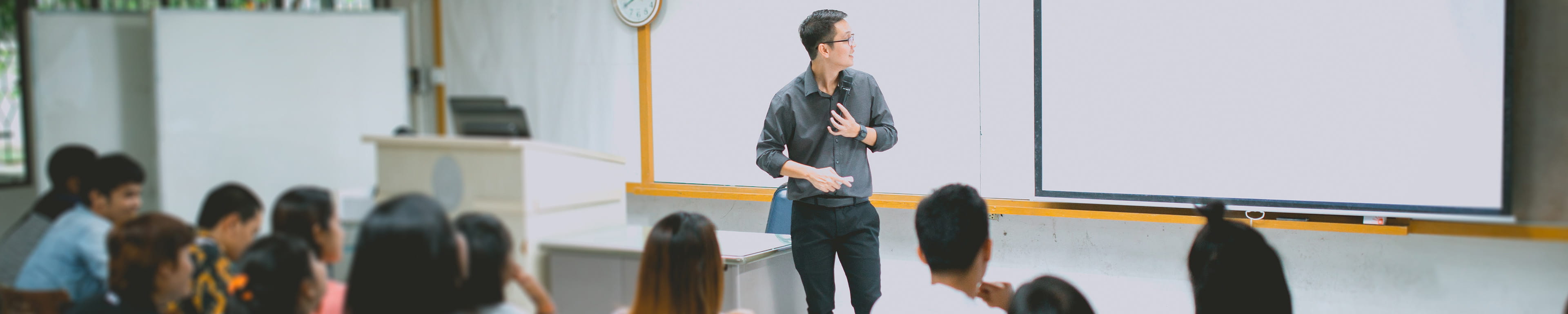 A man teaching a class full of students from the front of the class and whiteboard area. 