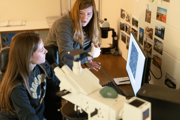 A student and instructor look at a computer screen displaying microscopic images in a lab, with a microscope in the foreground.