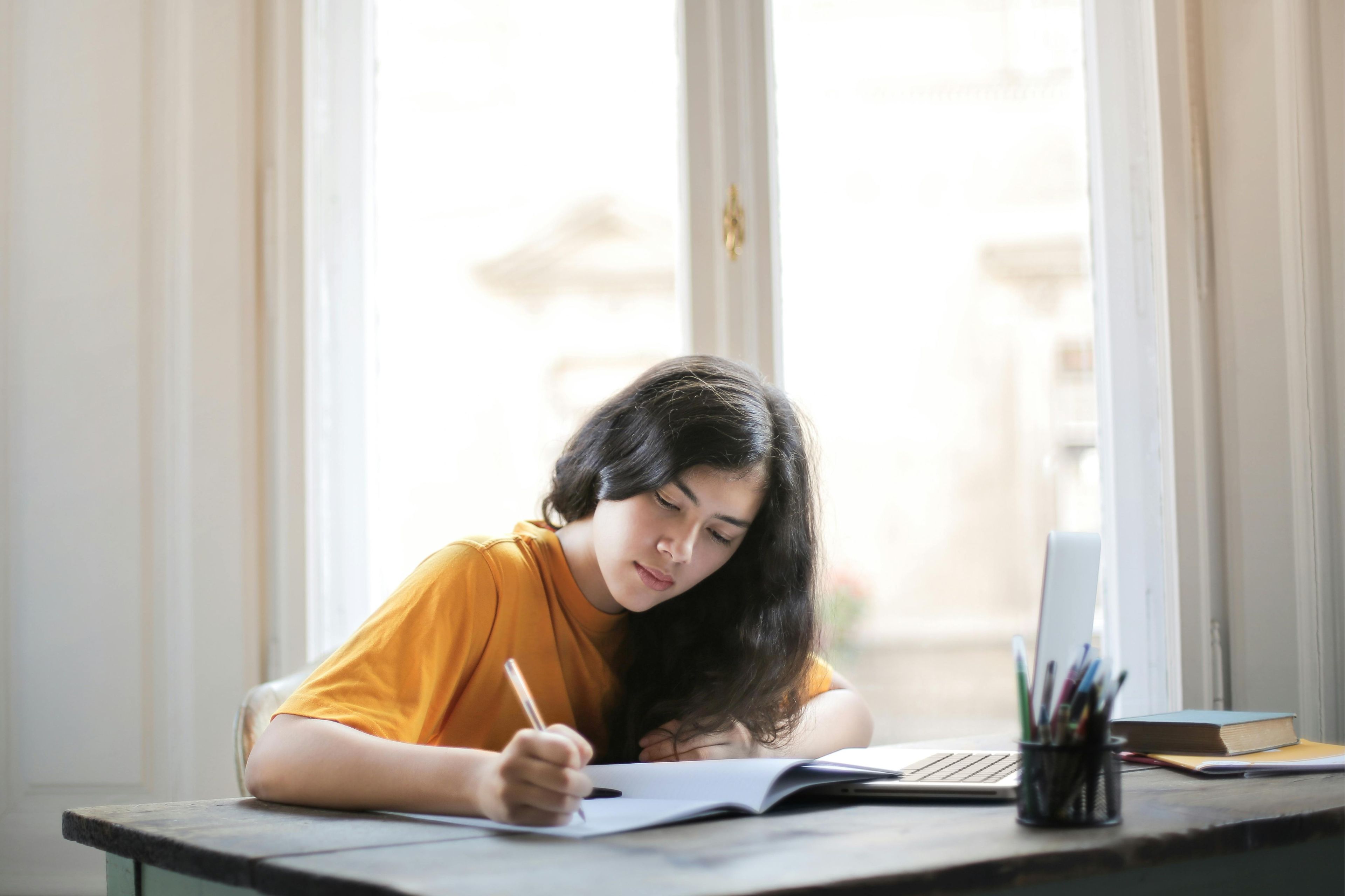 Young person sitting at a table with a pen in hand writing in a notebook.