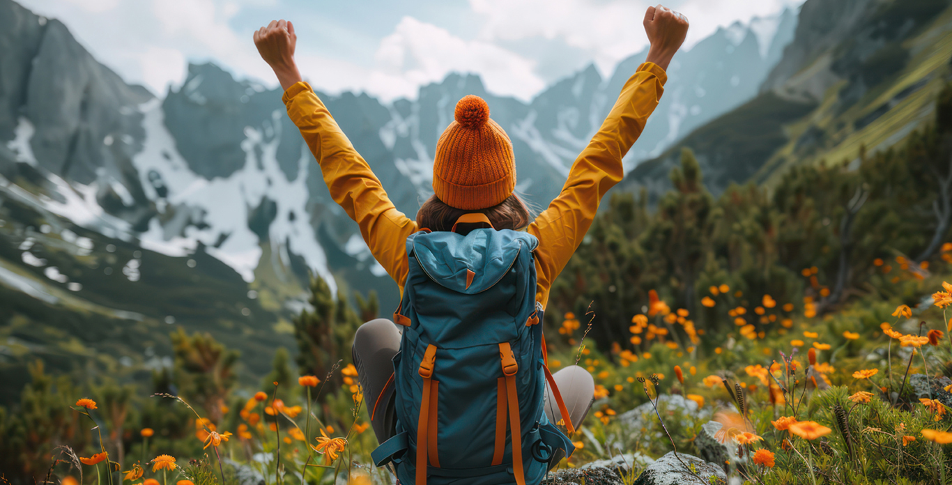 Person in gold jacket and hat and blue back pack with their arm stretched out in victory with a mountain scene in front of tee;e