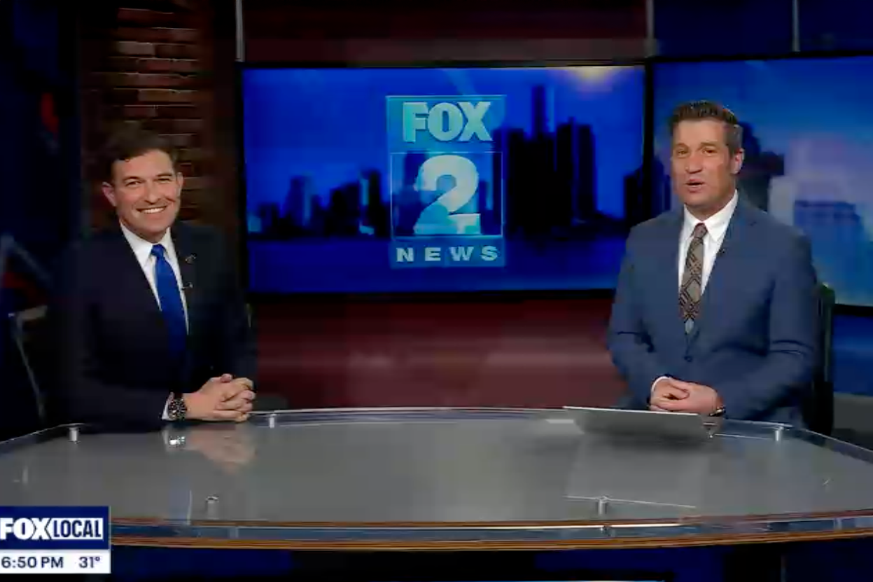 President Kavalhuna sits at a news desk with a news anchor.