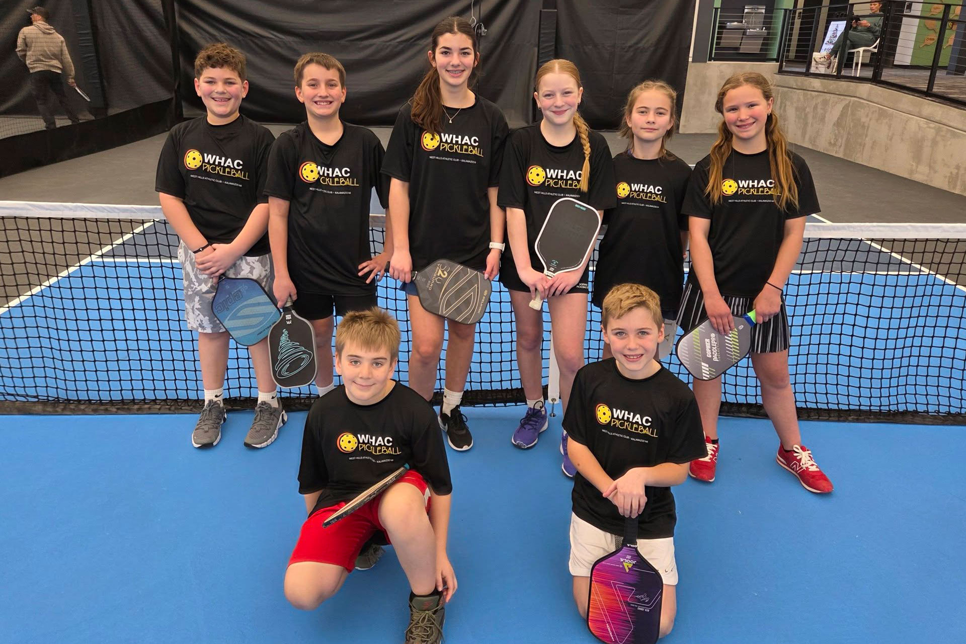 Eight boys and girls in front of the net for a youth pickleball team photo.