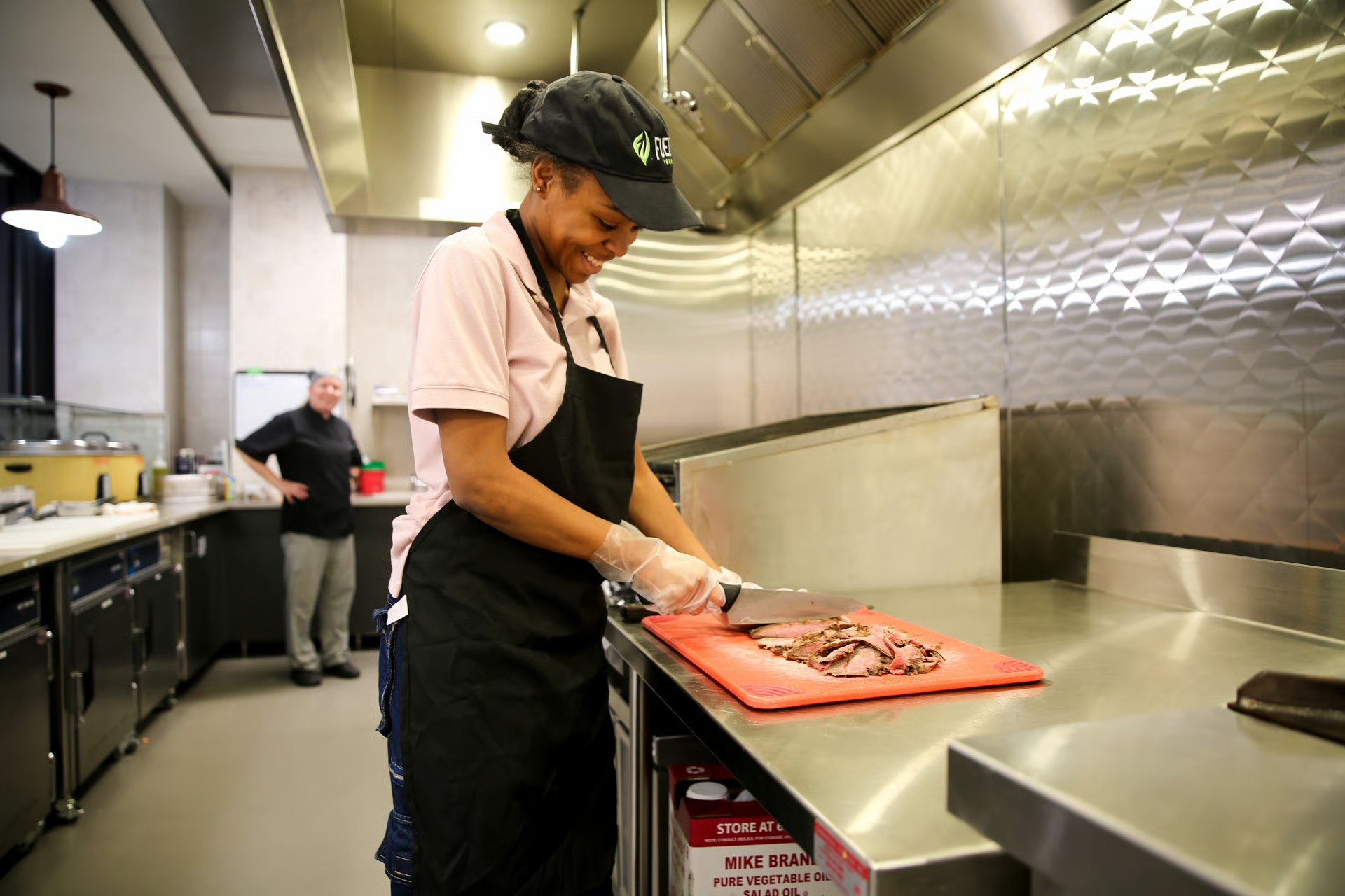 An employee wearing a black hat and apron chops meat on a cutting board in an industrial kitchen.