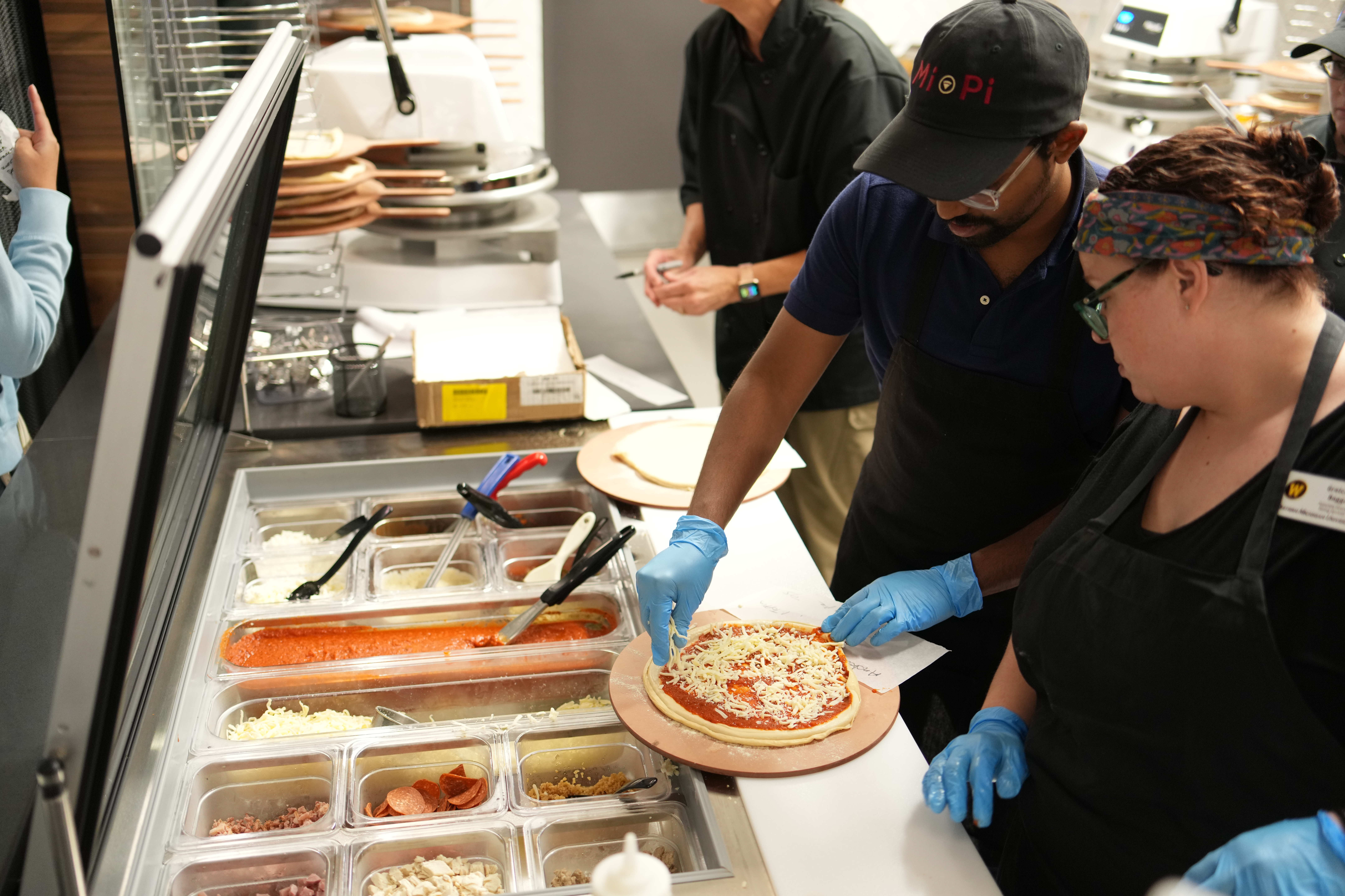A My Pie employee standing in front of ingredient trays sprinkling cheese on a pizza.