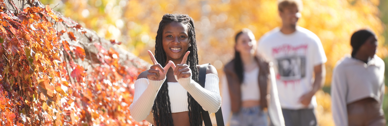 Female student on campus in the fall holding up her hands to form the letter w.