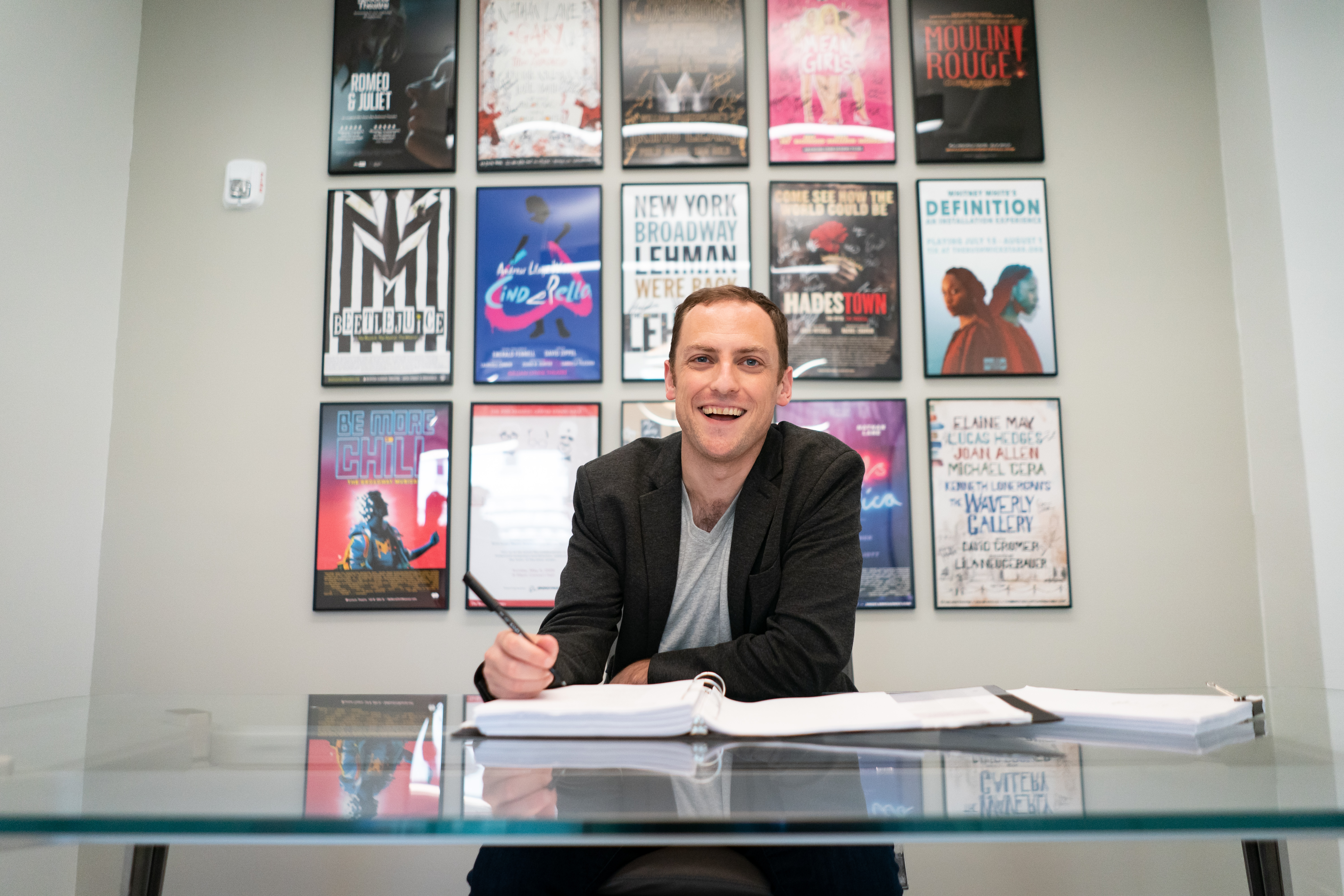 A man site at a desk with Broadway show posters framed behind him. 