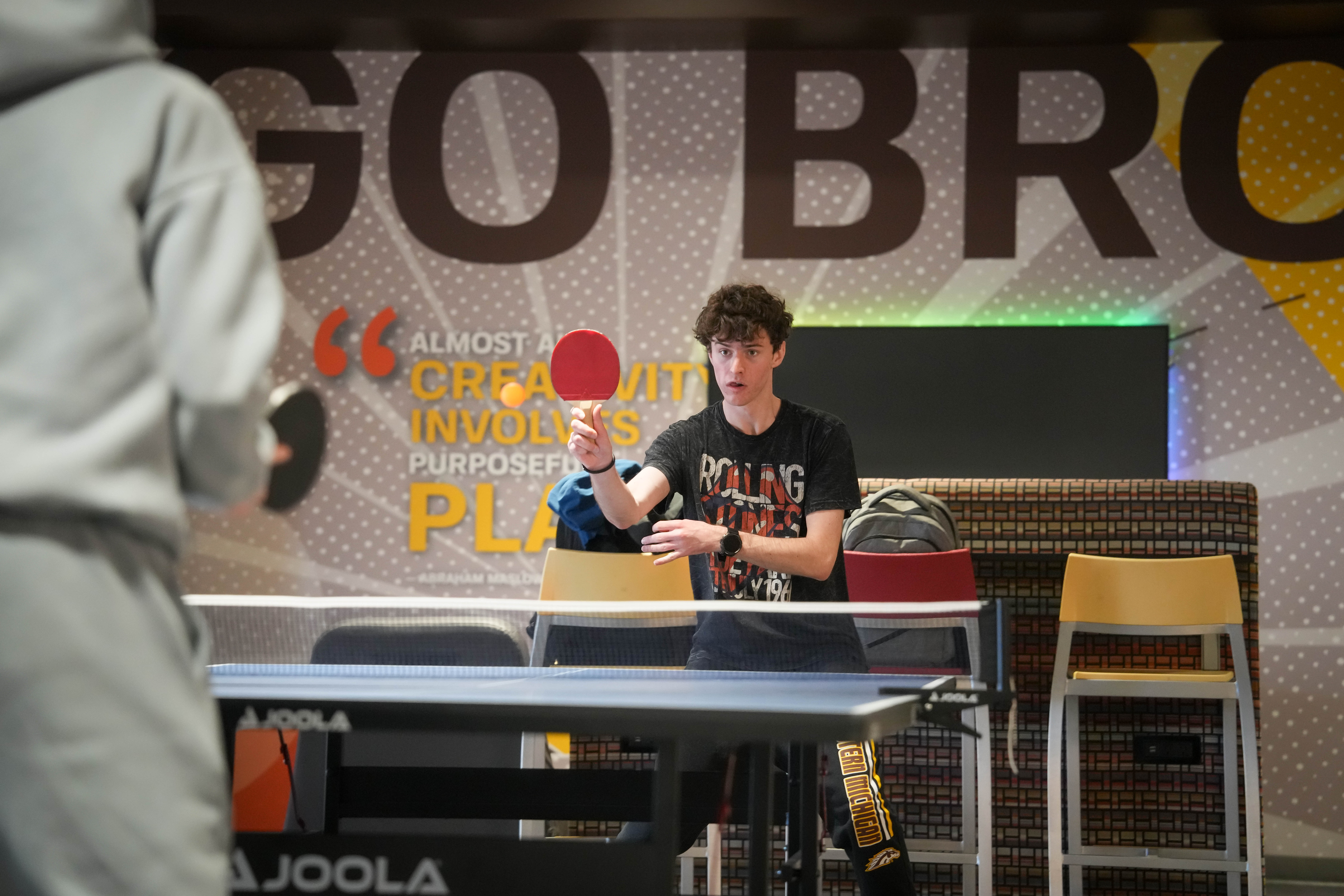 A student playing table tennis holds a red ping pong paddle in front of a large brown, gold, and orange Go Broncos mural.