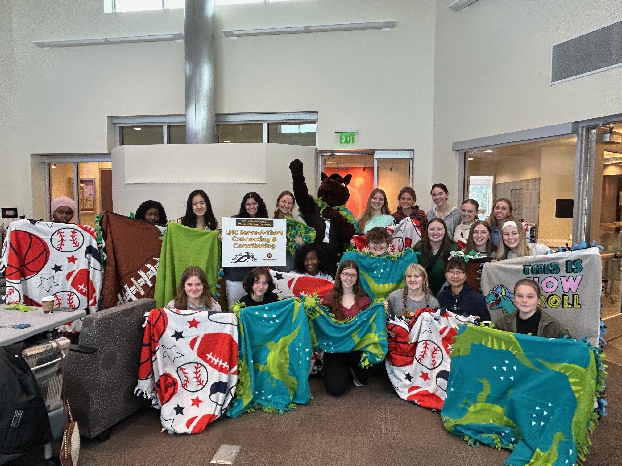 A large group of honors students are gathered in the honors college lounge holding up blankets they made for a local charity.