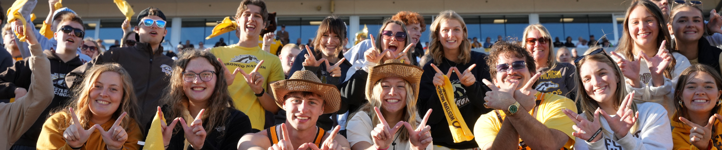 WMU students cheer and make the “W” hand sign while gathered in the stands at a Western Michigan University football game.