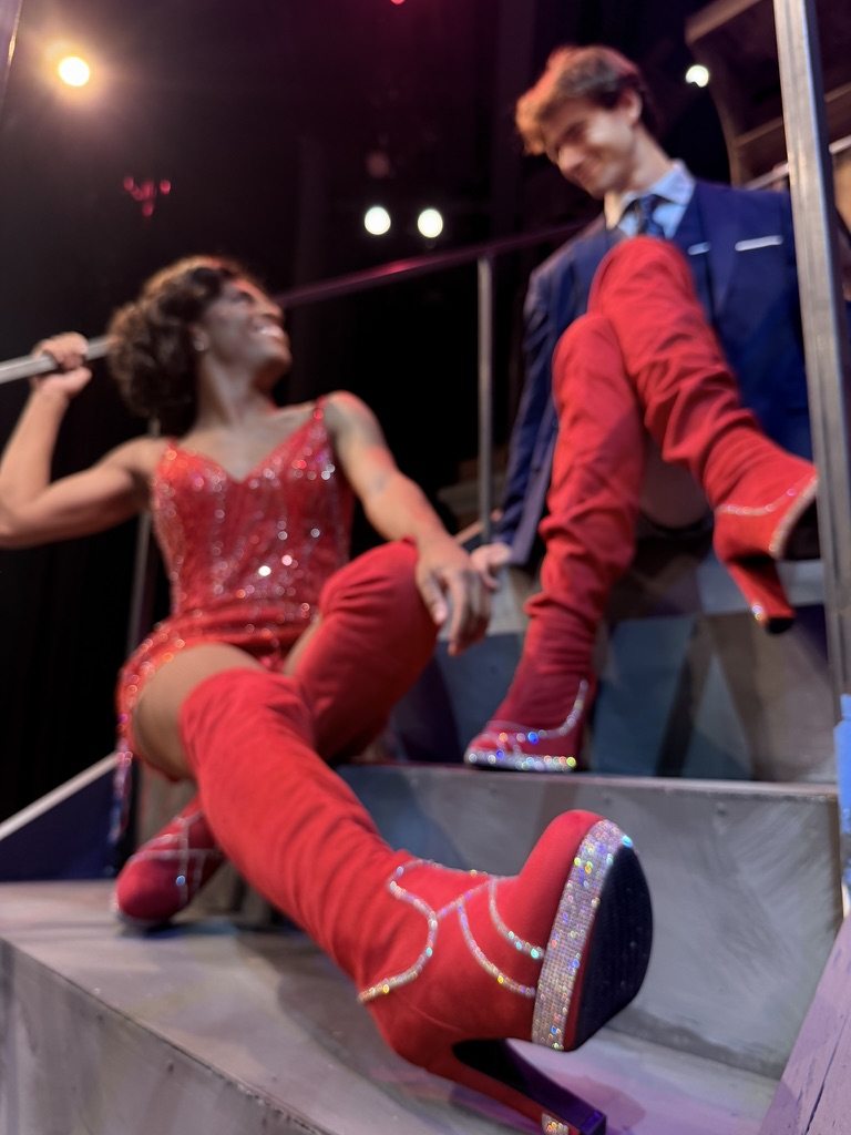 Man in suit and drag queen in red dress, both in red boots, look at each other on stairs