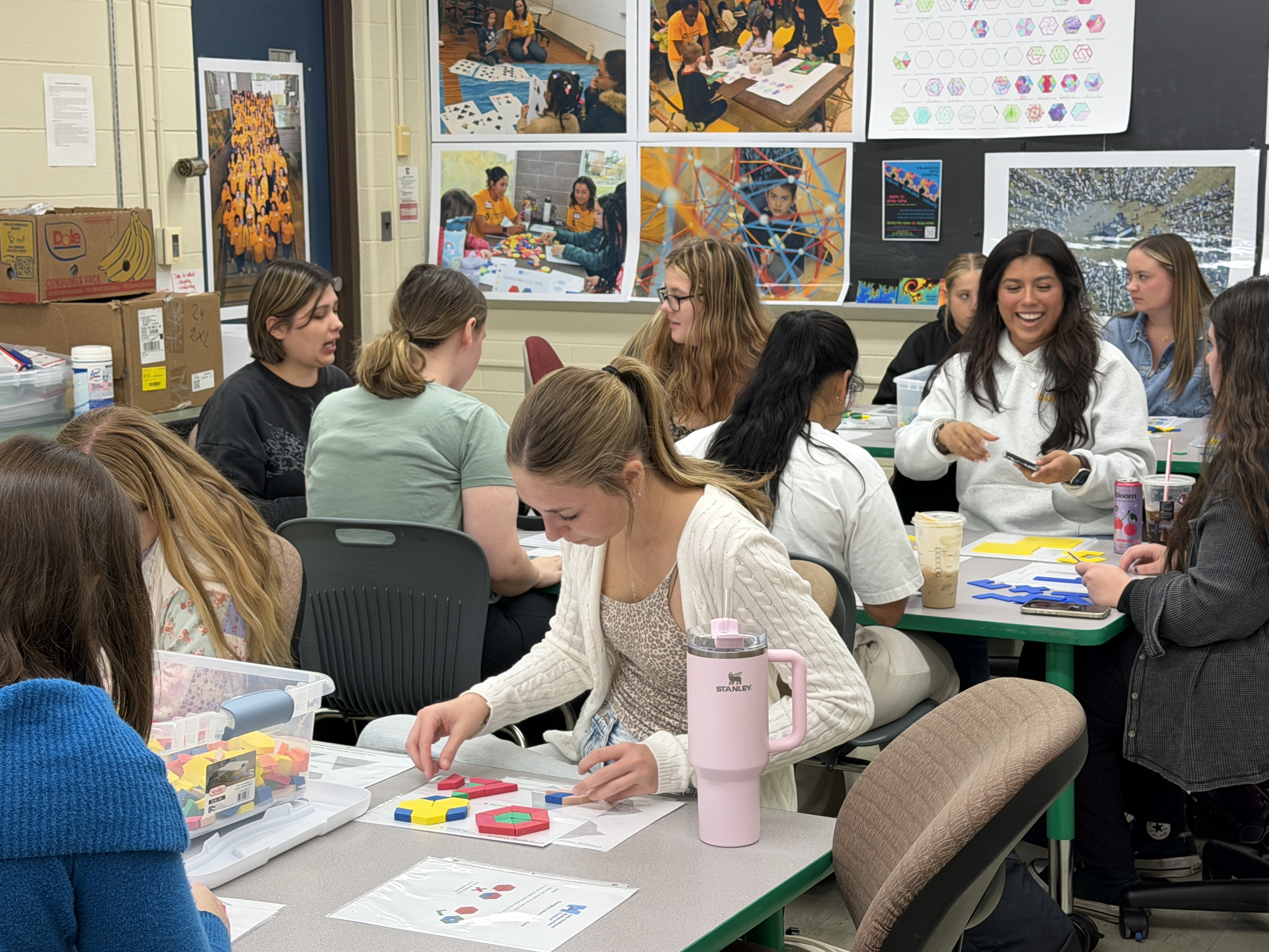 Multiple students sitting at tables using math manipulatives, like colored shapes to create. 