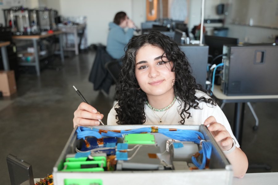 A student stands in a classroom and smiles at the camera. She holds a pen and is opening a drawer filled with supplies.