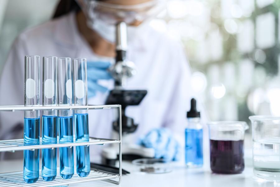 Scientific test tubes filled with blue liquid stand up in the foreground while a scientist looks into a microscope in the background.