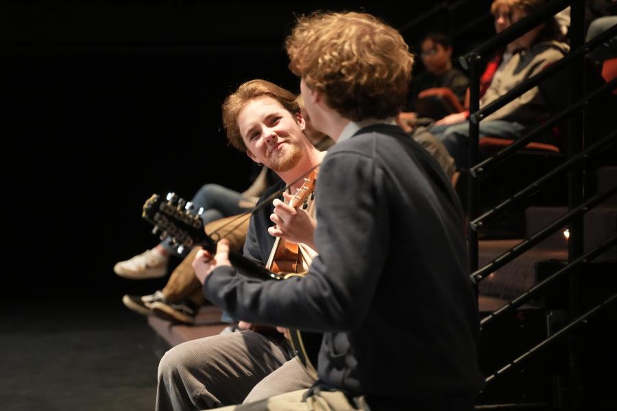 Two actors sitting with small guitars, playing and smiling together.