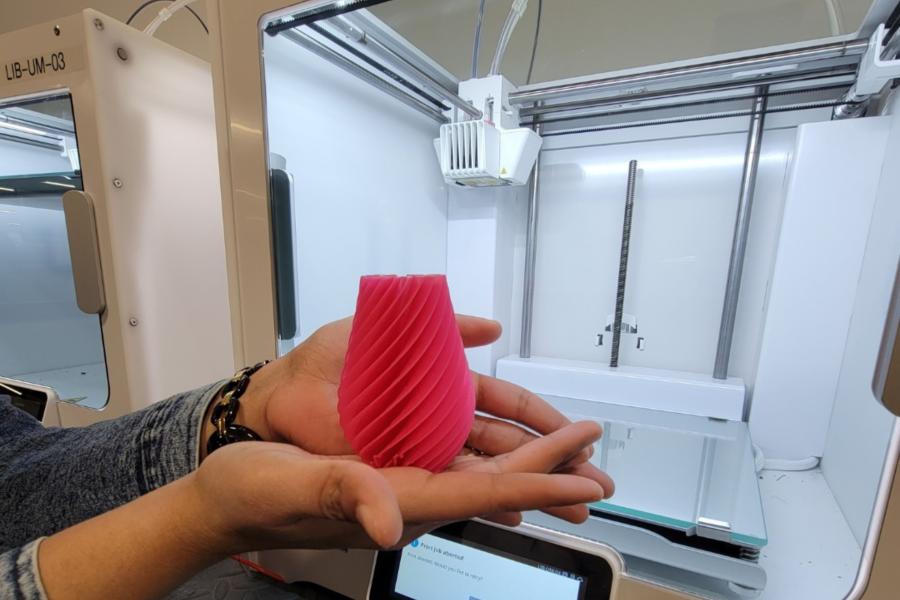 Hands holding a red 3d printed object in front of a 3d printer.