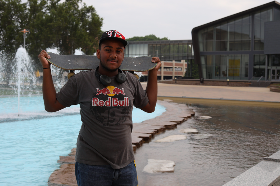 Yoyo Rodriguez Vargas holds a skateboard across his shoulders in front of Miller Fountain.