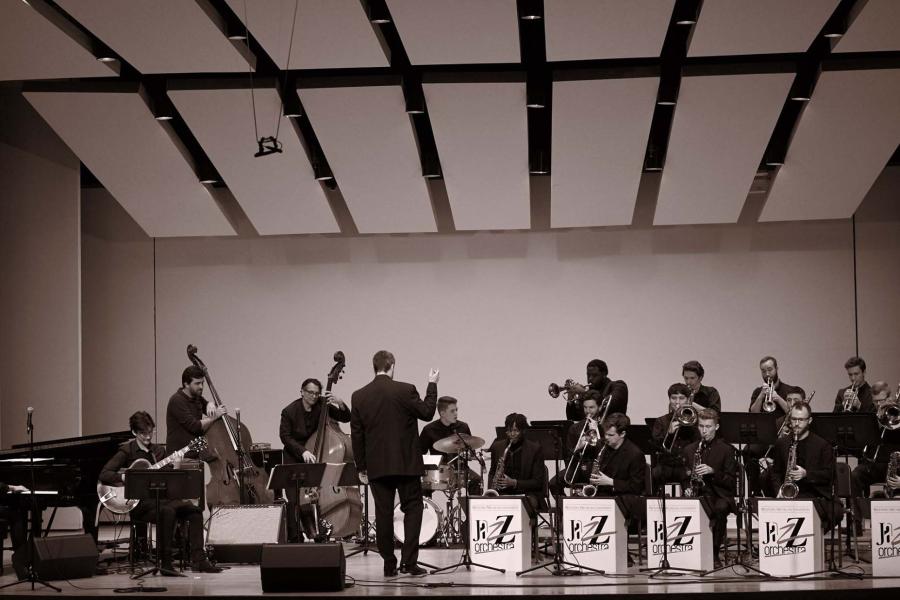 A jazz big band performs on stage. Featuring a conductor, piano, guitar, two basses, drum set, trumpets, trombones, and saxophones.
