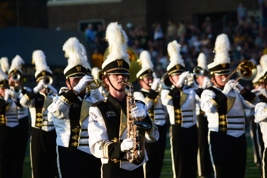 Members of a matching band performing on a football field. Their uniforms are white and gold. 