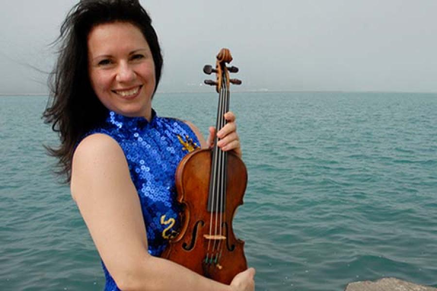 A smiling woman holding a violin stands in front of a large body of water.