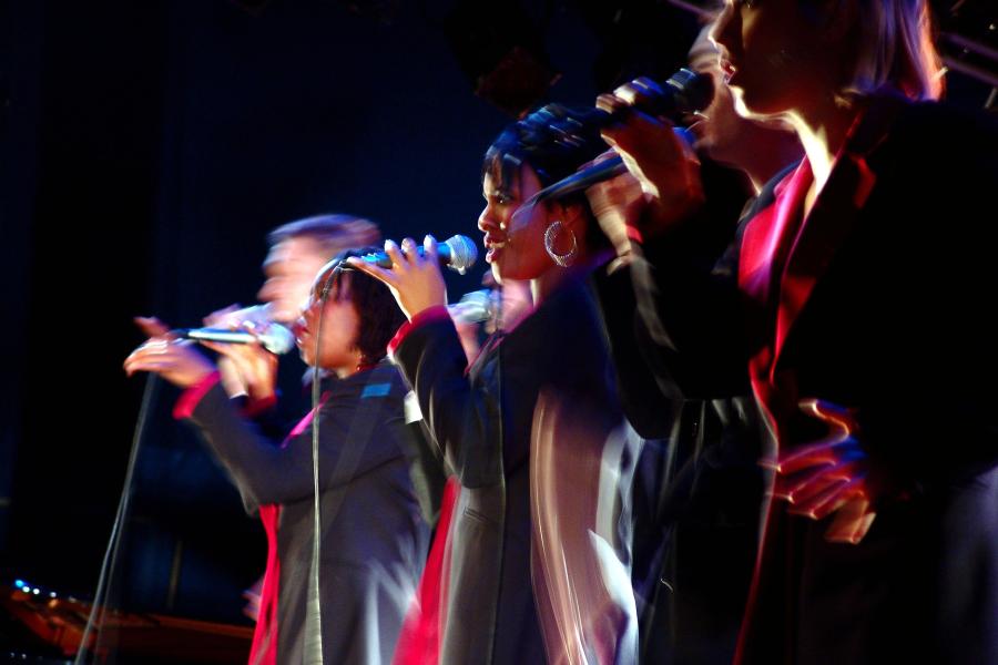 Stylized photo of multiple singers wearing red, singing with microphones. The image is artistically blurred, reflecting the motion of the performers.