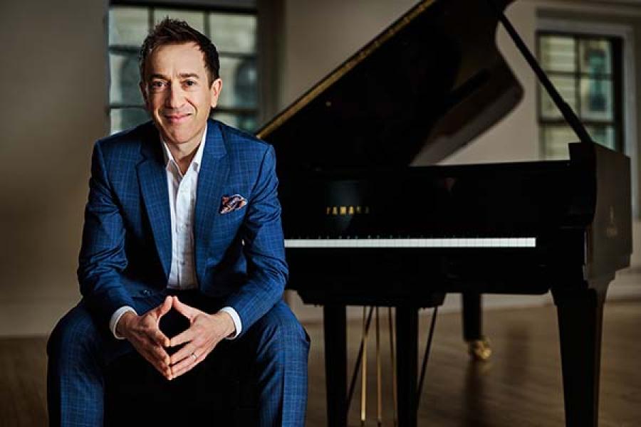A smiling man in a blue checkered suit sits in front of a grand piano.