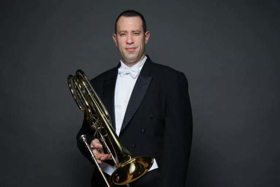 A smiling man in a black tuxedo holding a bass trombone.