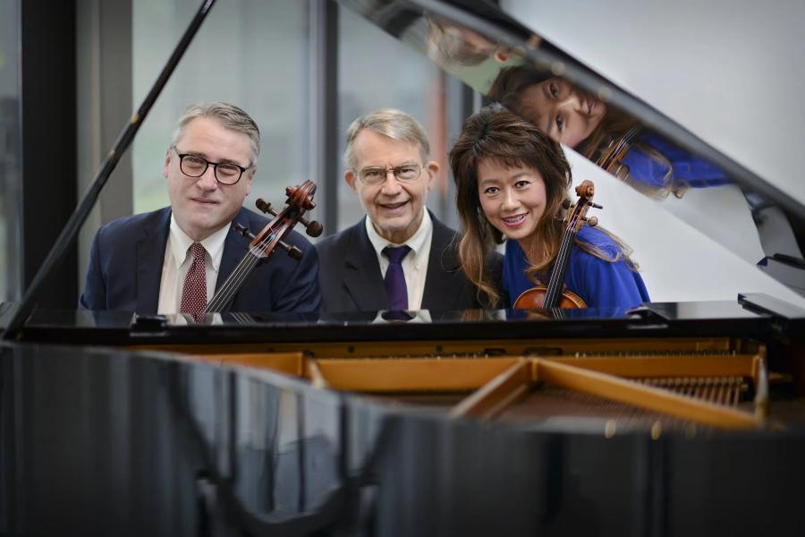 Three smiling musicians sitting behind a piano. The musician on the left is holding a cello, and the musician on the right is holding a violin.