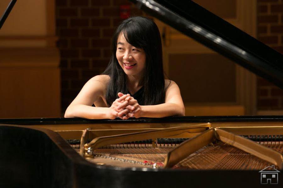 A smiling woman with long dark hair sits behind a piano.