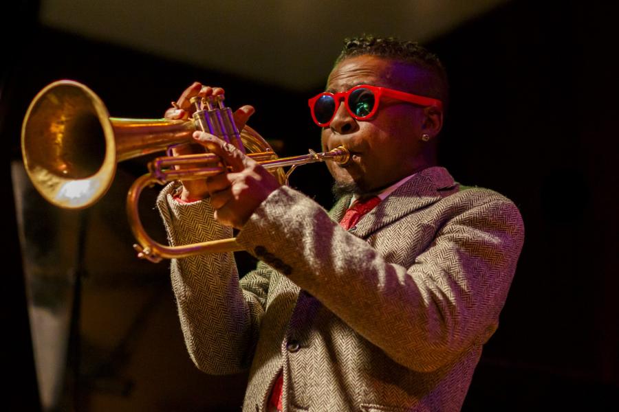A man wearing a tweed suit and red sunglasses plays the flugelhorn.