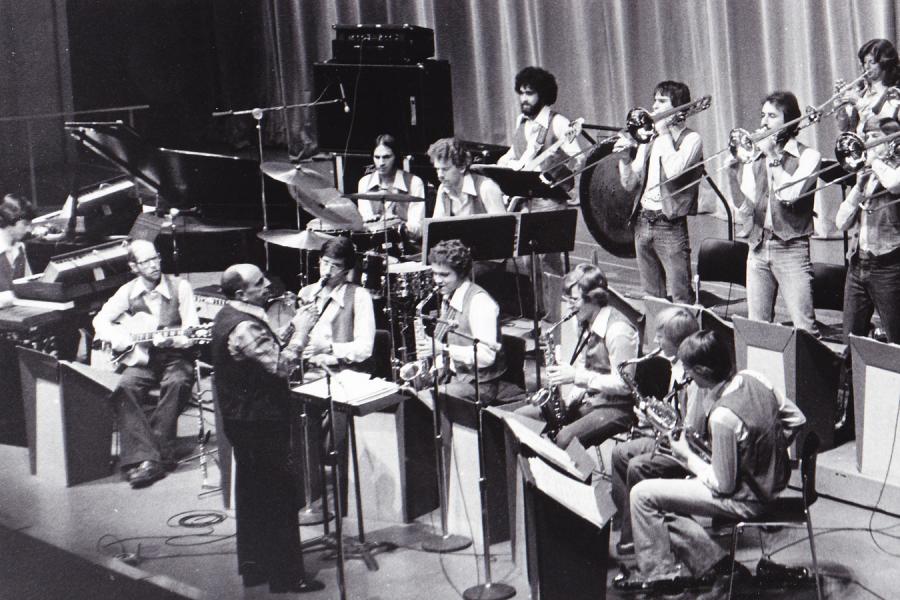 Black and white photo of a jazz ensemble performing. Saxophones, trombones, drums, electric guitar, electric bass, keyboards, and conductor are all on stage.