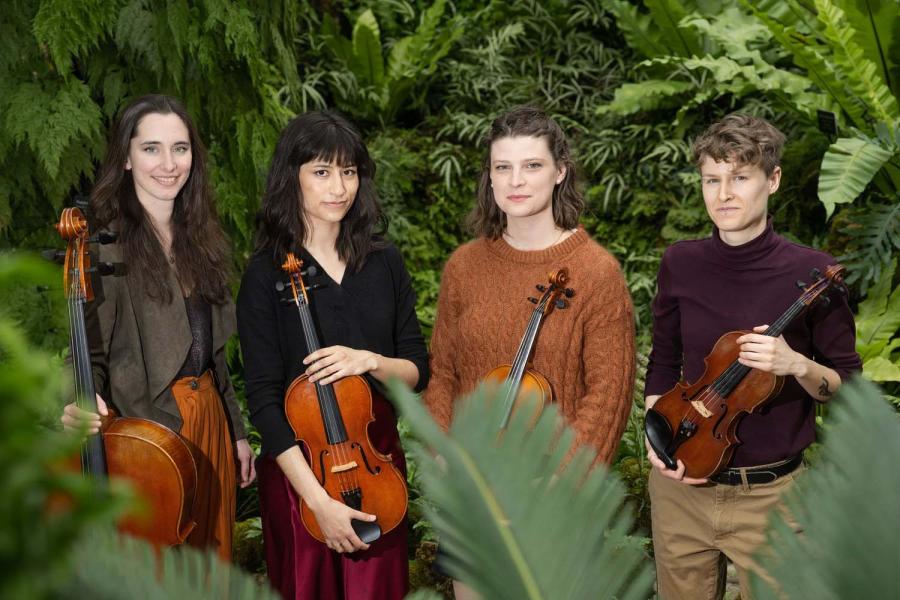 Four musicians (holding a cello, viola, and two violins, respectively) stand in a forested area.
