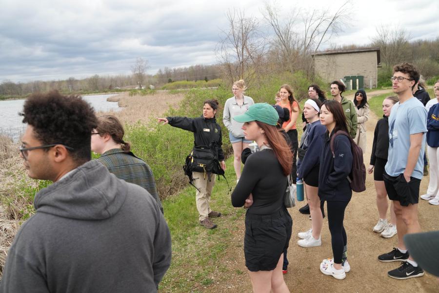 A group of college students stands on a dirt path near a lake and marshland, listening to a guide who is pointing toward the water at the Wolf Lake State Fish Hatchery.