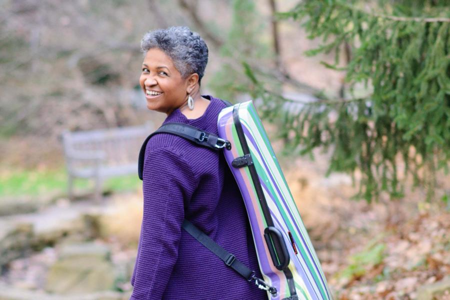 A smiling woman in a purple shirt looking over her shoulder. On her back she is wearing a colorful viola case.