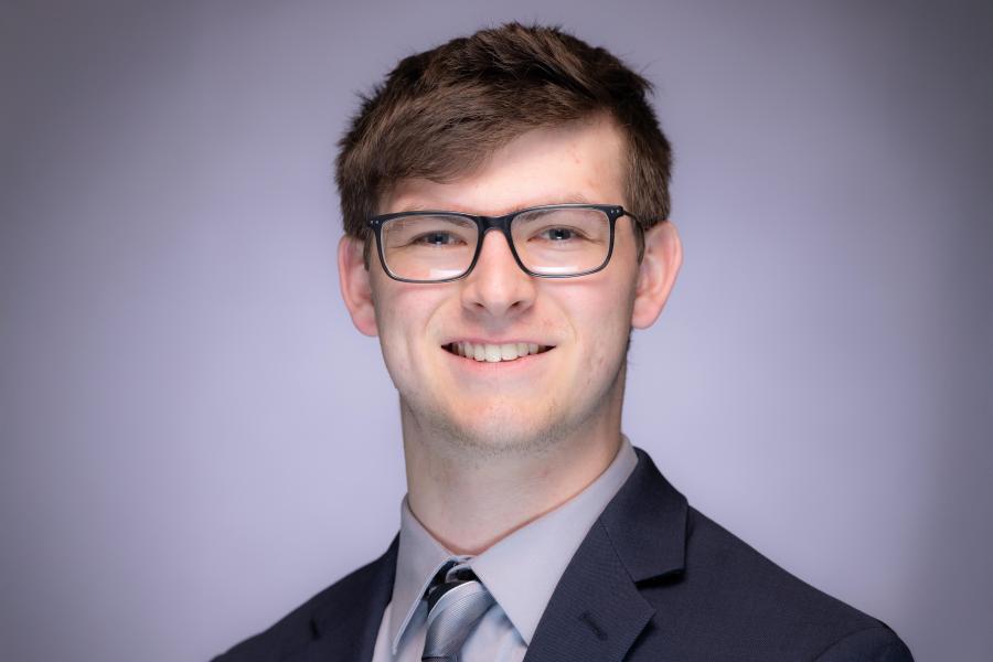 Headshot of Kevin Wright in a black suit with a grey stiped tie on a grey background