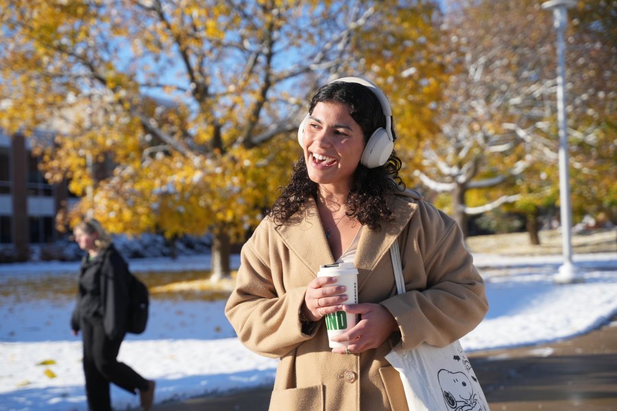a student with headphones and a coat on smiles at something outside of the frame. She's holding a cup and a tote bag. 