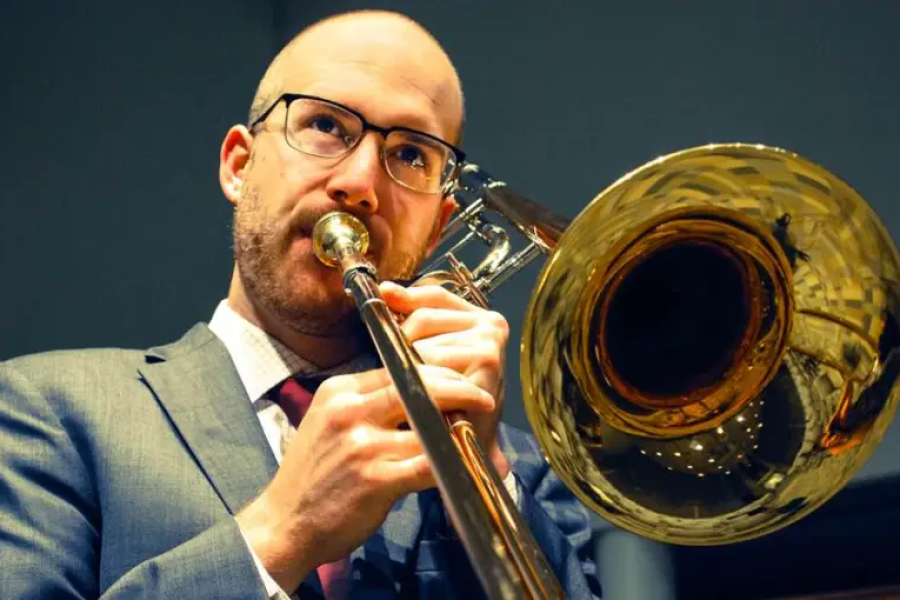 A man wearing a suit and glasses plays a trombone.