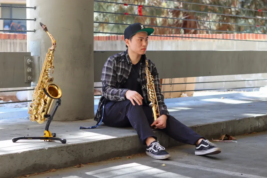 A man in a plaid shirt and a baseball cap sits on a curb outdoors holding a soprano saxophone. An alto saxophone sits on a stand to the left.