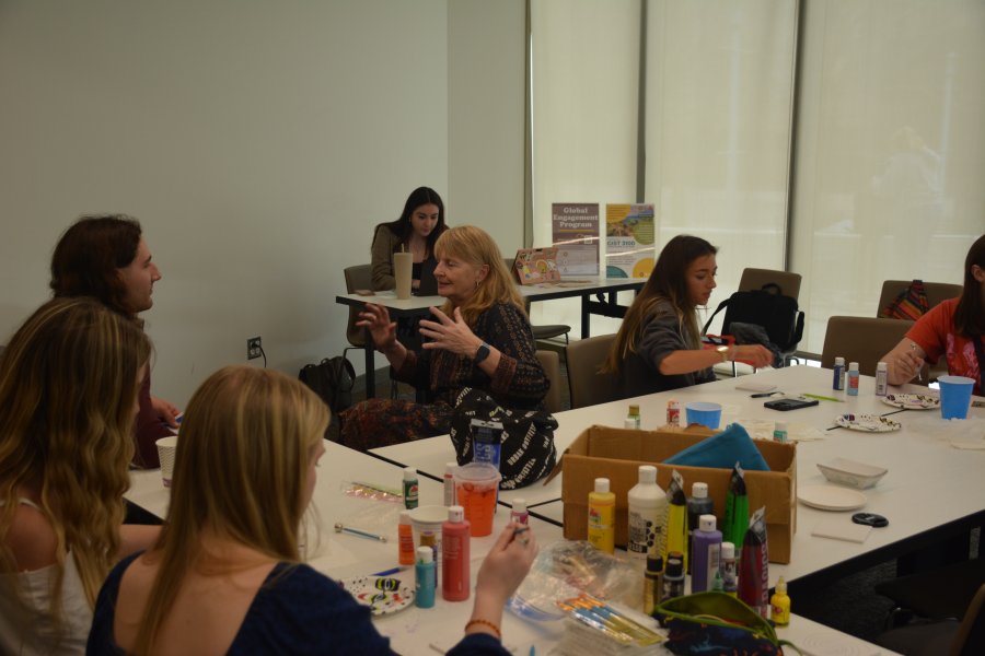A photo of 4 students sitting at a table and crafting with paint, while a professor engages in conversation with a fifth student.