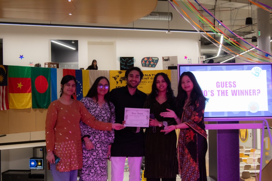 International students pose with an award at International Bazaar in the Student Center. Flags from many countries hang behind them.