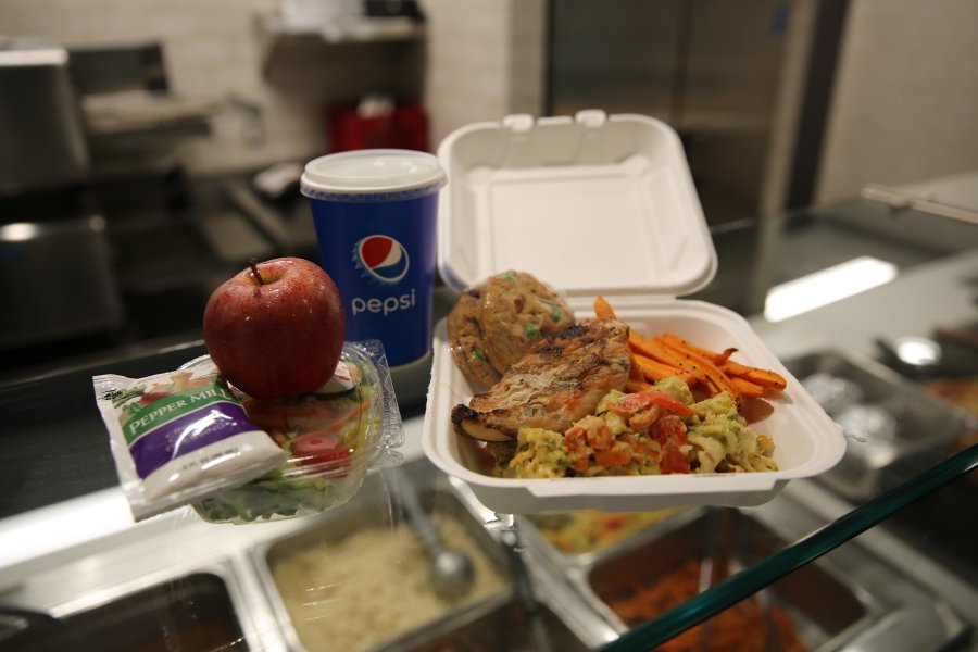 A selection of hot carryout food items in a styrofoam container next to an apple and a Pepsi cup.