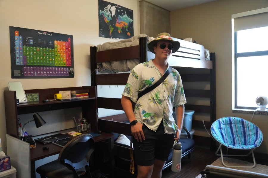 A student wearing a hat and sunglasses standing beside his desk and lofted bed.