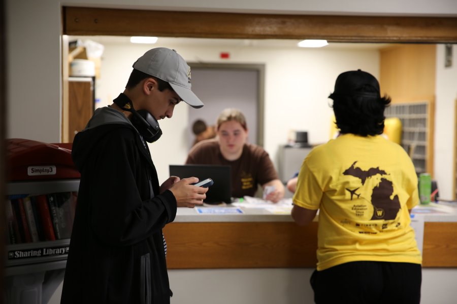 A student employee sitting at a residence hall service desk helps two students.