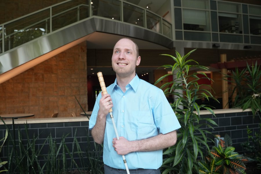 An undergraduate with a guide cane stands in an atrium with tropical plants.