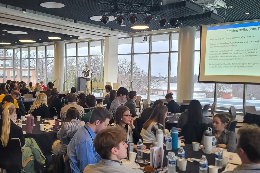 A crowd of people sitting around tables. They're listening to a presentation where the speaker has a powerpoint on the screen. 