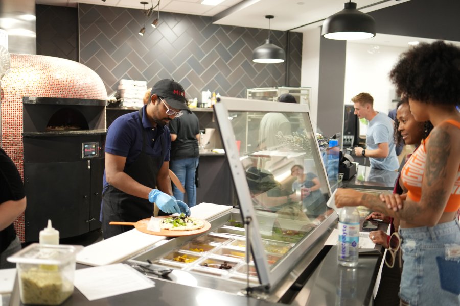 A student employee in a MiPi hat and apron assembles a pizza for a patron standing in line.