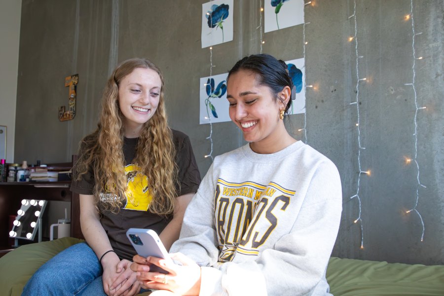 Two roommates wearing WMU-branded clothing sit together in their decorated campus room.