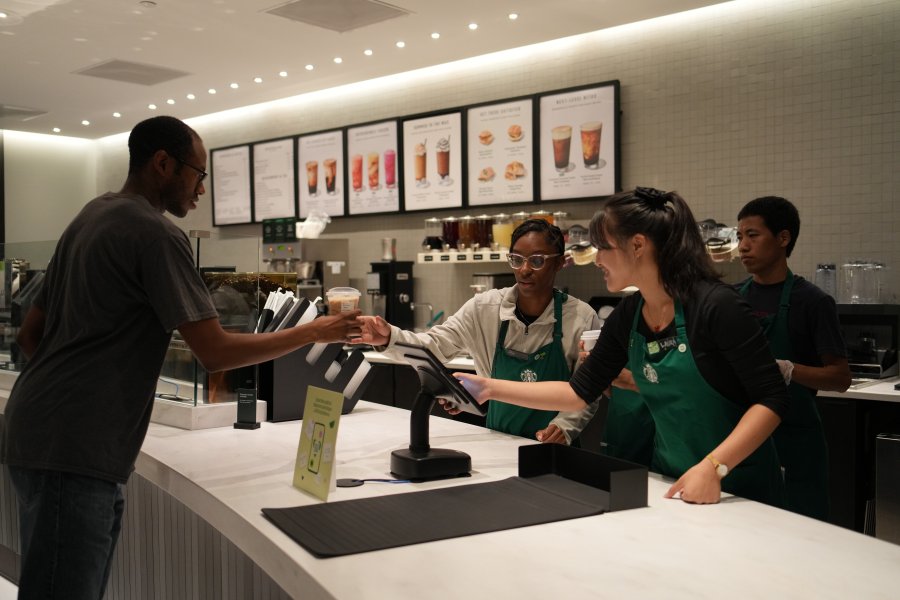 Two student Starbucks employees serve a to-go coffee to a patron.