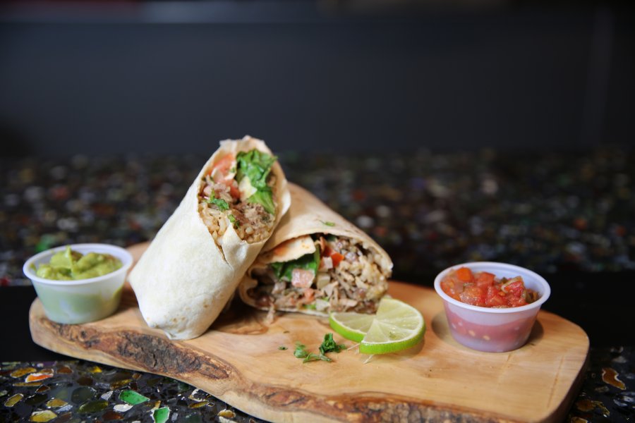 A burrito cut in half sitting on a decorative wooden plank between small cups of salsa and guacamole.