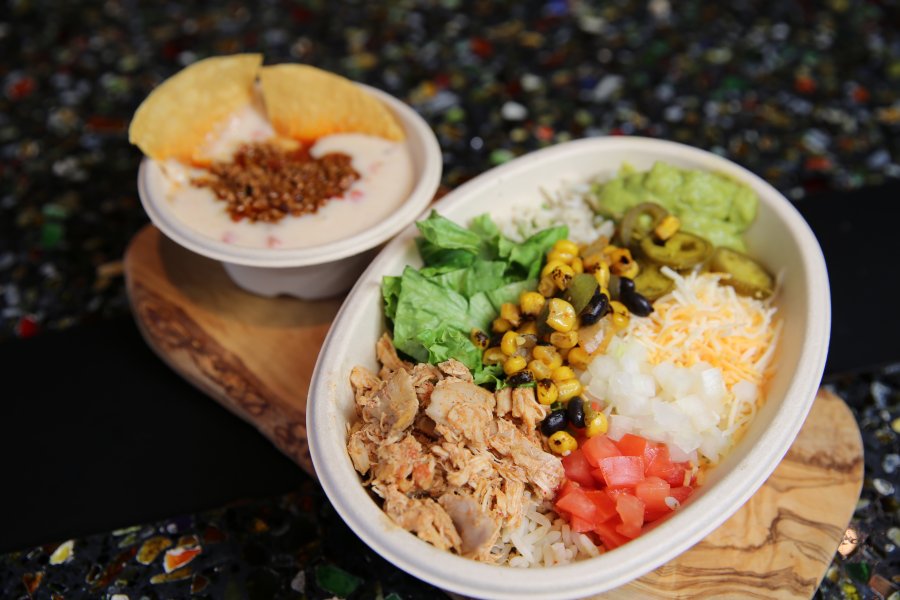 A burrito bowl sits on a decorative wooden plank beside a bowl of chips and queso.
